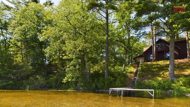 A wooden cabin with a deck, surrounded by dense forest and a clear lake.