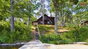 A wooden dock extending into a clear stream, surrounded by lush greenery and a cabin in the background.