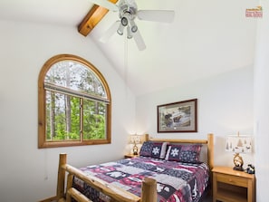 Une chambre dotée d’une grande fenêtre en arc de cercle, d’un ventilateur de plafond, d’un lit recouvert d’une courtepointe à motifs, de tables de chevet avec des lampes et d’un tableau encadré au mur.