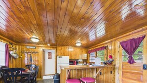 A wooden cabin interior with a stone-tiled floor, a dining area with chairs, a kitchen with a refrigerator, and a bar area with stools.