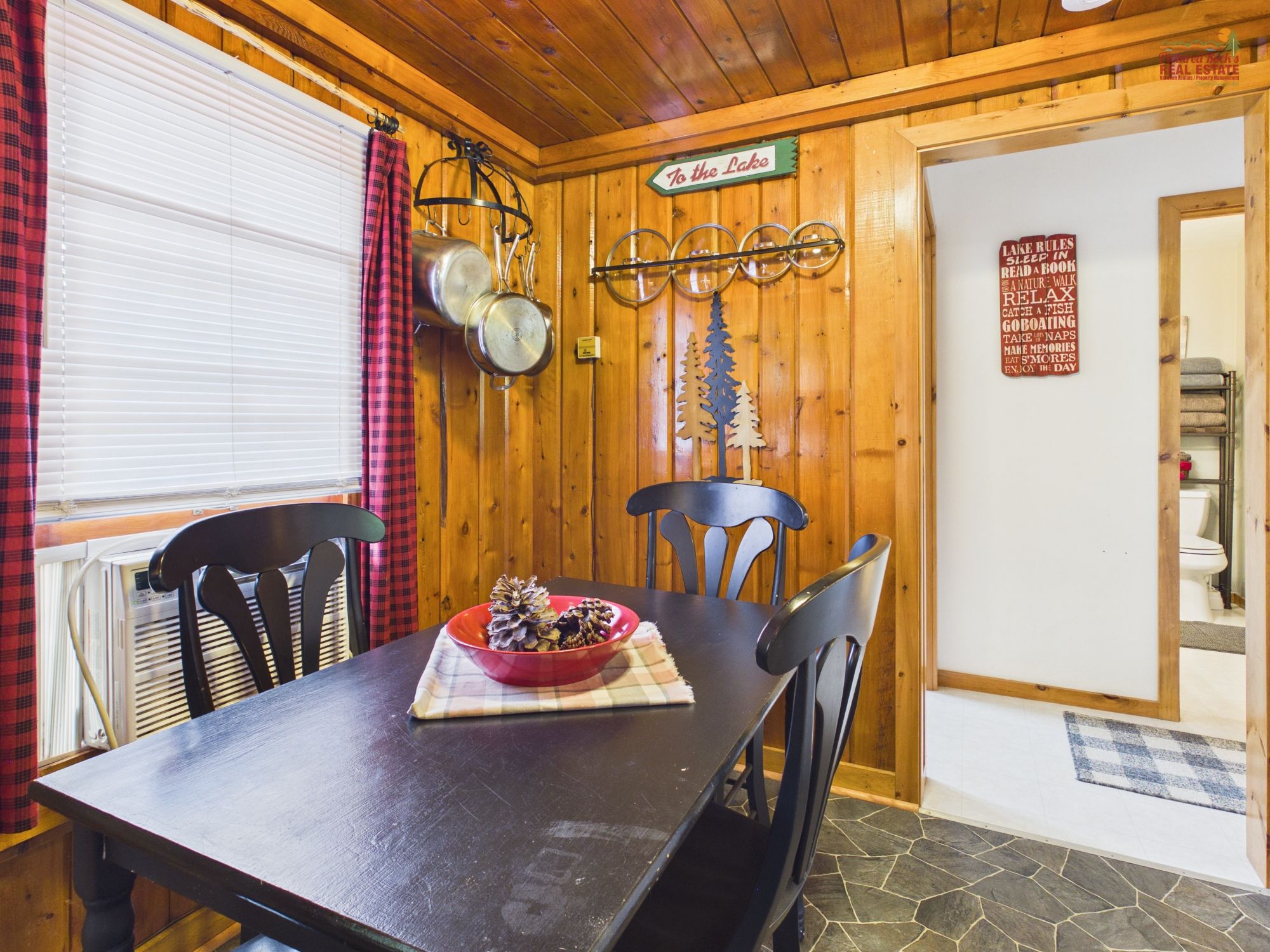 Interior de una cabaña de madera con una mesa de comedor, sillas, un tazón de comida, una ventana con persianas y una puerta con un letrero.