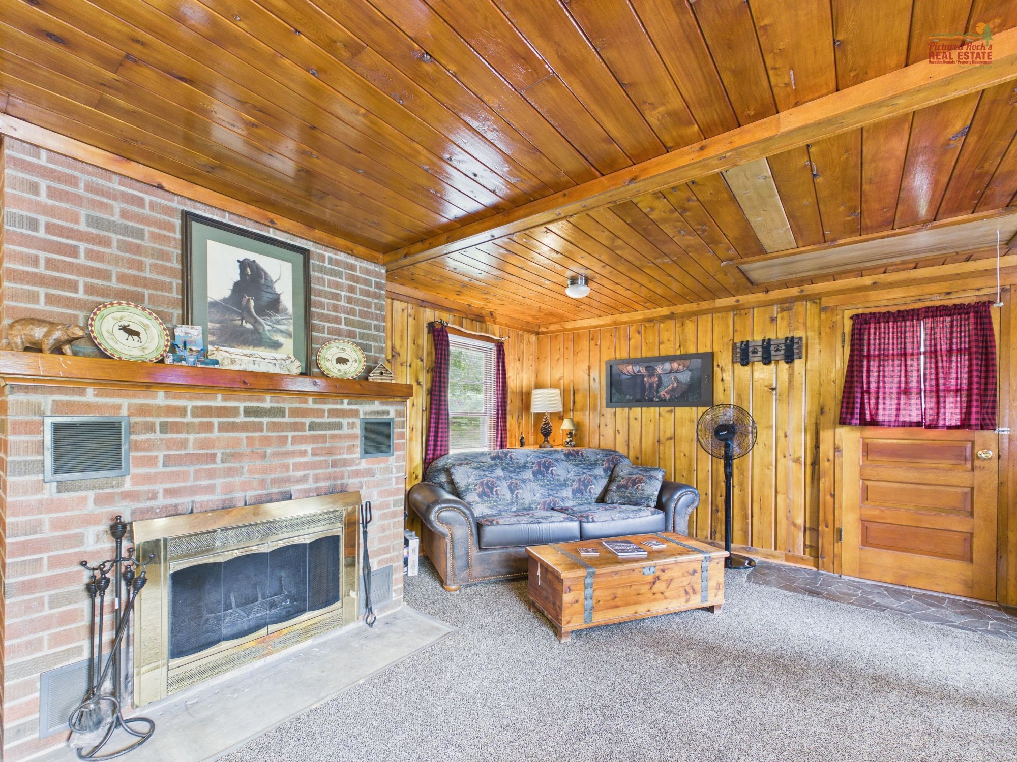 A cozy cabin interior with a fireplace, a sofa, a wooden chest, and a fan.