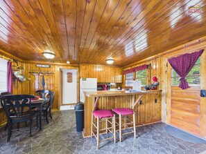 A wooden cabin interior with a stone-tiled floor, a dining area with chairs, a kitchen with a refrigerator, and a bar area with stools.