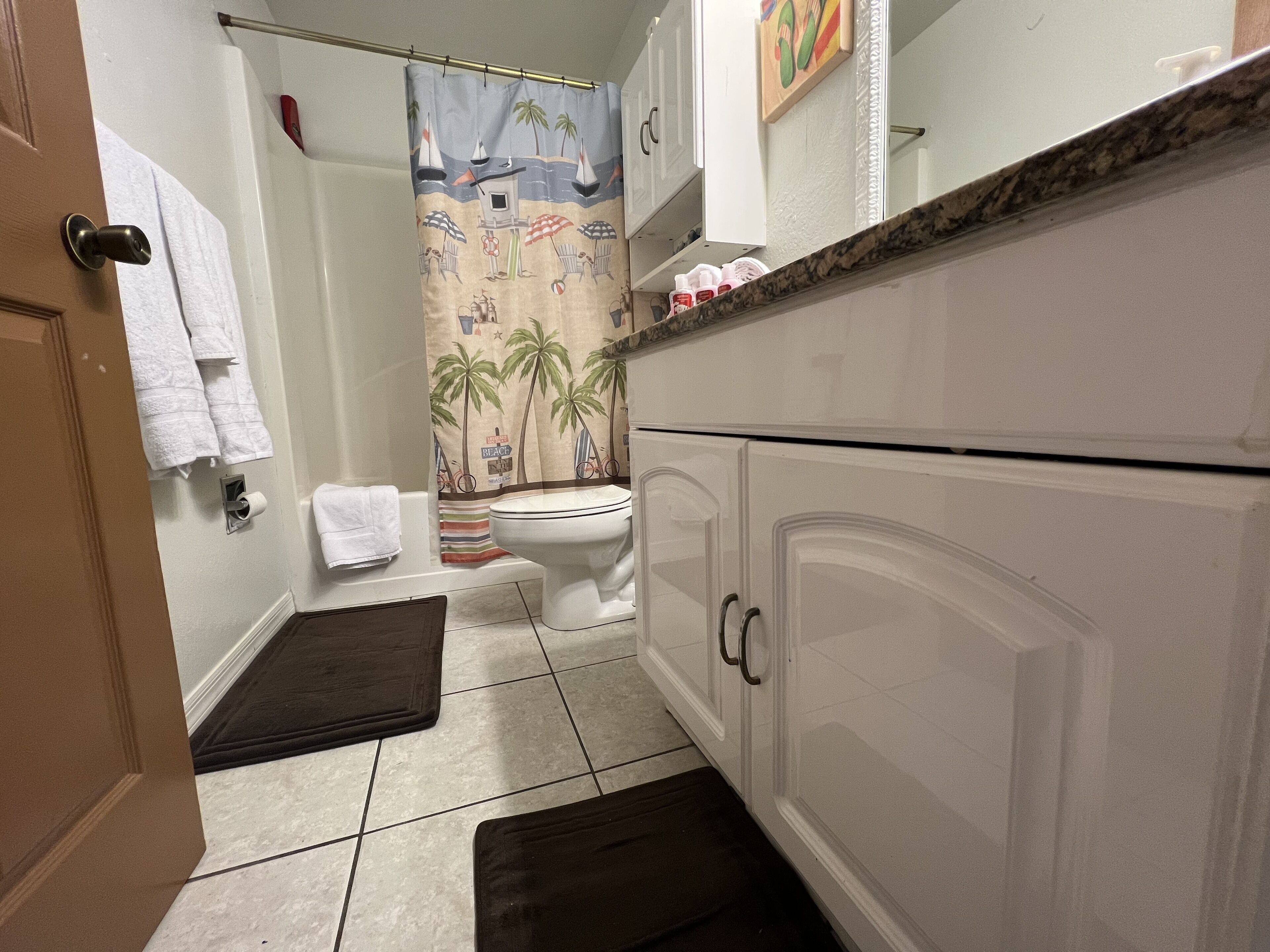Une salle de bain avec des toilettes, un rideau de douche à motifs de plage, des meubles blancs et un porte-serviettes.