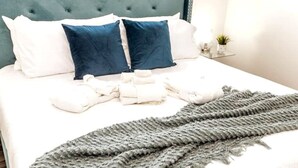 A neatly made bed with a grey knitted throw, white and blue pillows, and a tufted headboard. - Chic Suite - Long Stays - King Bed  - Fireplace &WiFi
 (Edmonton)