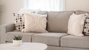 Ein wohnzimmer in neutralen Farbtönen mit einem beigen Sofa, zwei gemusterten Zierkissen, einer kleinen Topfpflanze und einem runden Couchtisch.