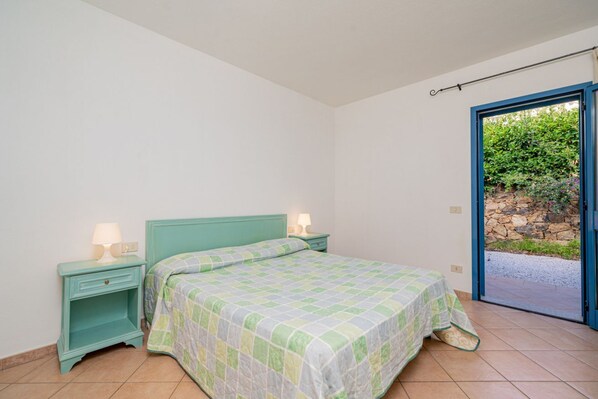 Un dormitorio con una cama, dos mesitas de noche con lámparas, una ventana con vistas a la vegetación y una puerta que da al exterior.