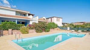 A swimming pool with a tiled deck, surrounded by a white building with a tiled roof and a garden with various plants. - All have their own entrance and are located in four-family villas.Elegantly furnished, they have a small garden with furnished covered veranda or furnished covered terrace.Composed of living room with kitchenette and sofa, bedroom with double bed, room w (Monte Petrosu)