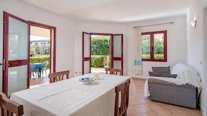 A dining area with a table set for two, a sofa, and a view of a garden through open doors. - All have their own entrance and are located in four-family villas.Elegantly furnished, they have a small garden with furnished covered veranda or furnished covered terrace.Composed of living room with kitchenette and sofa, bedroom with double bed, room w (Monte Petrosu)
