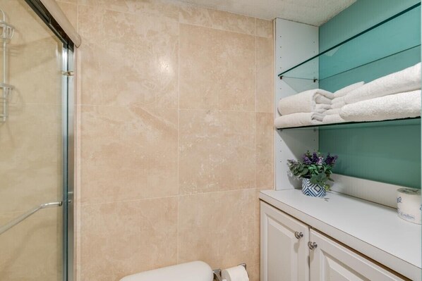 A bathroom with beige tiles, a glass shower enclosure, a white toilet, and a shelf with folded towels and a potted plant.
