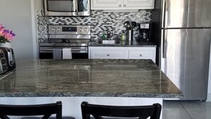 A modern kitchen with white cabinets, a granite countertop, a stainless steel refrigerator, and a ceiling fan.