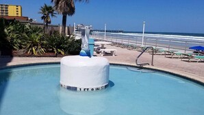 A swimming pool with a central fountain, surrounded by lounge chairs and palm trees, with a beach and ocean in the background.