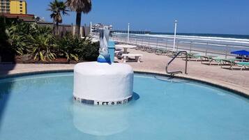 Une piscine dotée d’une fontaine centrale, entourée de chaises longues et de palmiers, avec une plage et l’océan en arrière-plan.