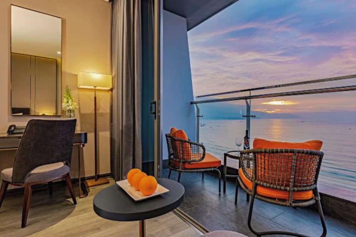 A balcony with wicker chairs, a small table, and a view of the sea at sunset.