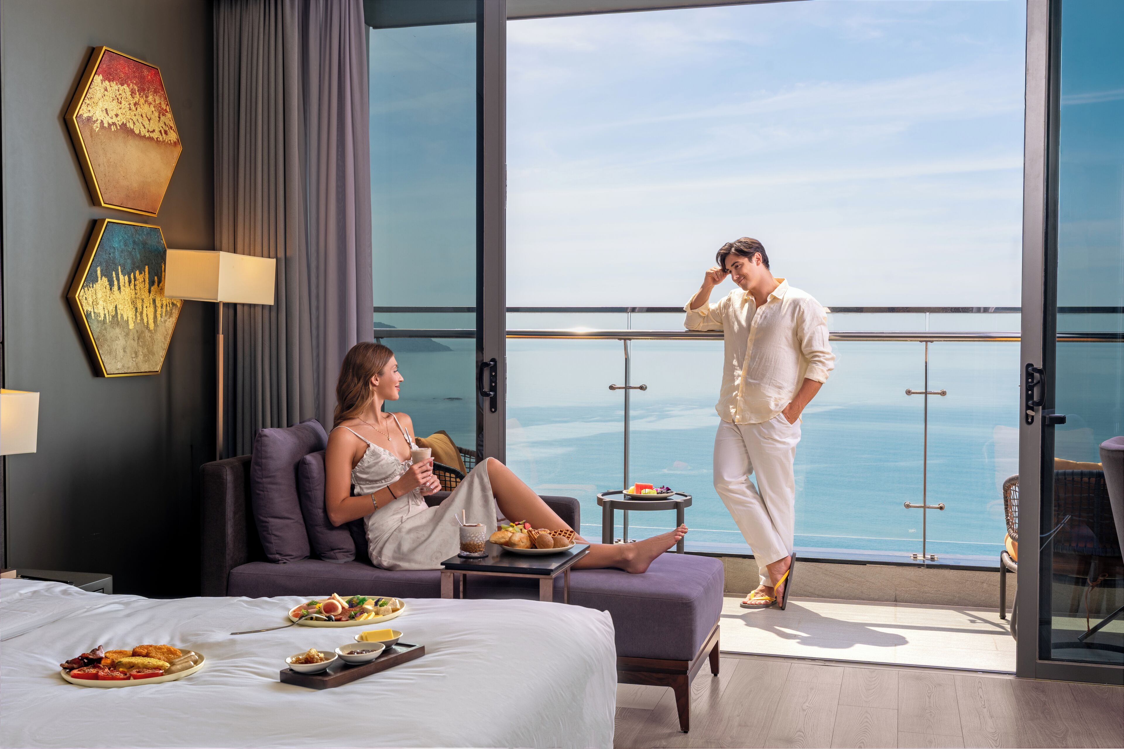 Habitación de hotel con cama, una mesa de comedor con desayuno, un balcón con vista al mar y un ventanal grande.