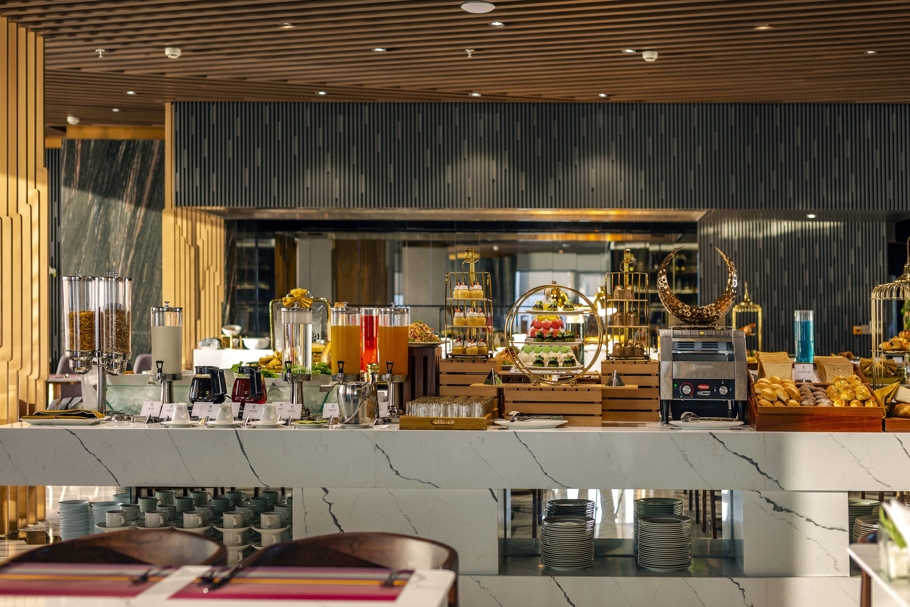 A buffet area with various food stations, including a coffee machine, fruit display, and a cake station.