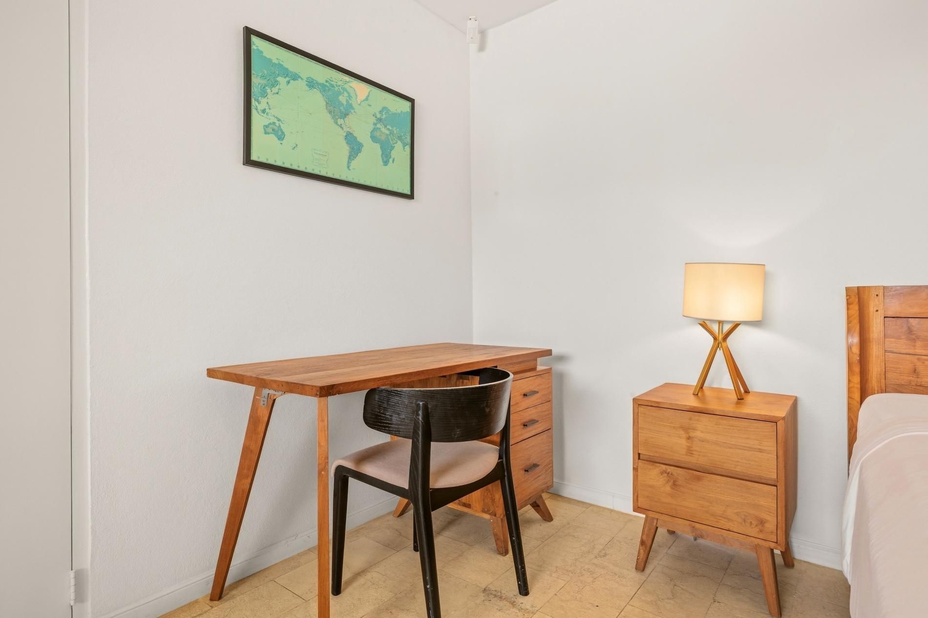 Une petite pièce comprenant un bureau en bois, une chaise, une table de chevet avec une lampe et un lit.