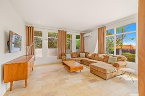 A spacious living room with a large sectional sofa, a wooden coffee table, and a side table. There are multiple windows providing natural light and views of the outdoors.