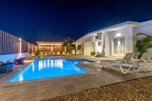 Una piscina moderna a l'aire lliure amb gandules, una zona de seients coberta i un porxo ben il·luminat.