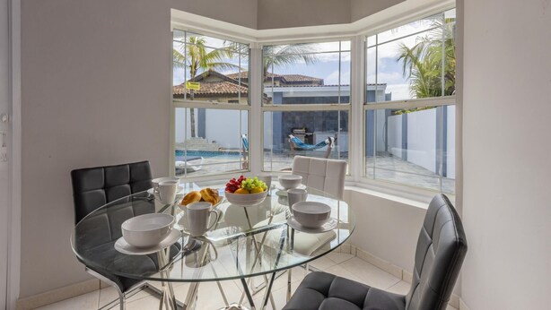 Un coin repas avec une table et des chaises en verre, donnant sur une piscine et des palmiers à l’extérieur.