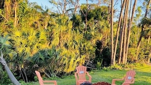 A grassy outdoor area with four red plastic chairs and a small table, surrounded by dense tropical vegetation and tall trees.