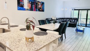 A modern kitchen with a granite countertop, a sink, and a dining area with a table and chairs. There is a black leather sofa and a large window in the background.