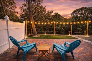 Pátio externo com duas cadeiras azuis e uma mesa de madeira, cercado por uma cerca branca e luzes de Natal.