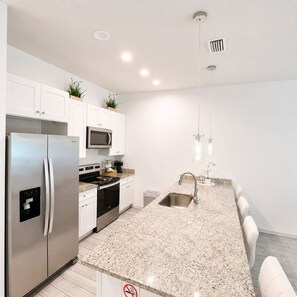 Une cuisine moderne avec des meubles blancs, des appareils en acier inoxydable, des plans de travail en granit et des luminaires suspendus.