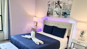 A neatly made bed with a blue quilt, a white headboard, and a white bedside table with a lamp.
