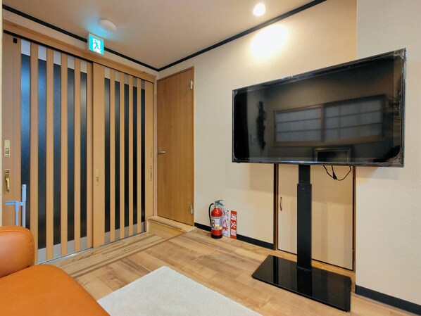 Una habitación con un televisor montado en la pared, una puerta de madera con listones verticales, un extintor y una alfombra blanca en el suelo.