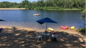 Un entorno junto al lago con una mesa y sillas bajo un paraguas azul, kayaks y un bote pequeño en la orilla.