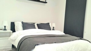 Een net opgemaakt bed met wit en grijs beddengoed, twee bedlampjes en een donkere, structuurrijke muur.