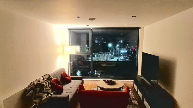 Een woonkamer met een bank, een rode stoel, een kleine tafel en een televisie. Er is een groot raam met uitzicht op een stadsbeeld 's nachts.