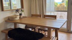 Un comedor con una mesa de madera, bancos, un felpudo negro, una cesta trenzada y un florero con flores.