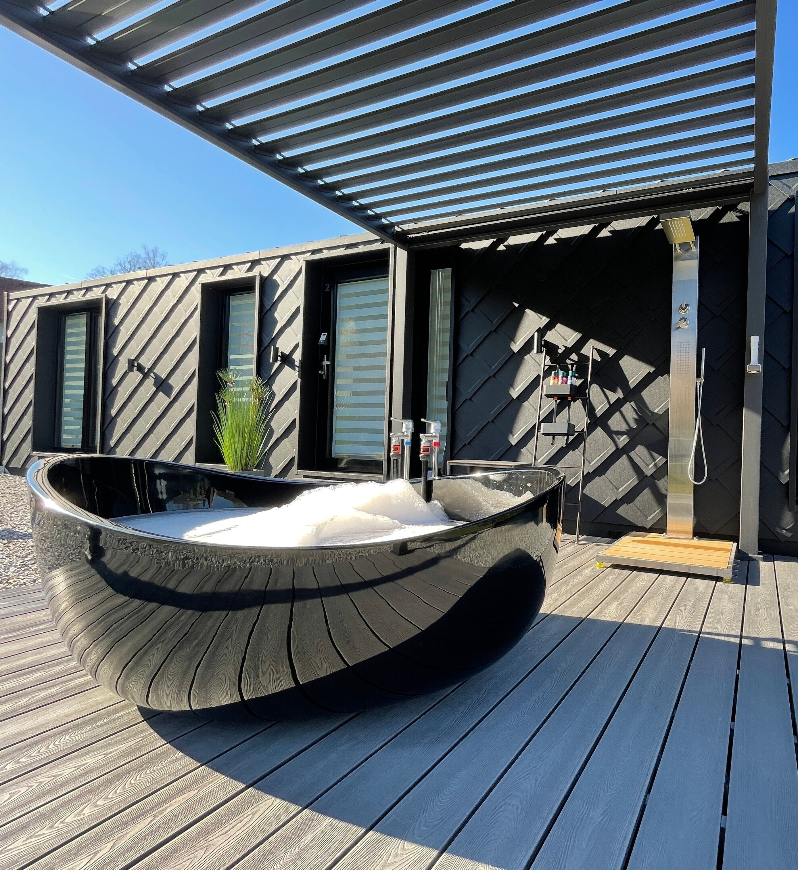 A modern outdoor hot tub with a wooden deck, a metal pergola overhead, and a black and grey patterned wall.