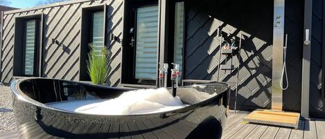 A modern outdoor hot tub with a wooden deck, a metal pergola overhead, and a black and grey patterned wall.