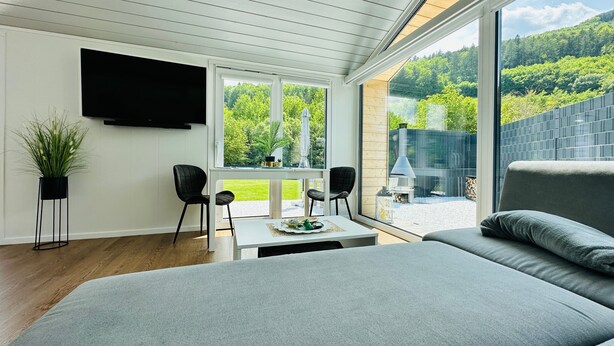 A modern room with a large bed, a TV mounted on the wall, and a view of a garden and mountains through large windows. - Holiday Home 'Voralpen Lodge - Premium' with Jacuzzi, Terrace & Wi-Fi - Adults Only (Traisen)