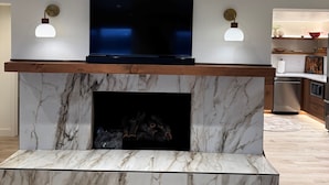 A modern living room with a marble fireplace, a flat-screen TV, and two wall-mounted lamps.