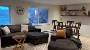 A living room with a sofa, ottoman, and a coffee table. There are windows with a view of trees and a body of water.