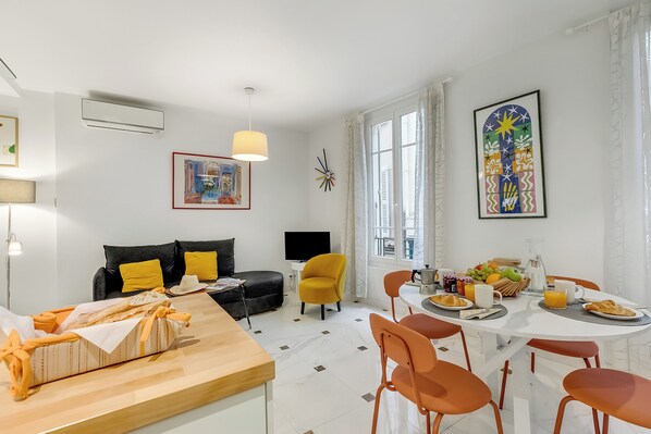 Ein modernes Wohnzimmer mit einer Couch, einem Esstisch, der für das Frühstück gedeckt ist, und einem großen Fenster mit Vorhängen.