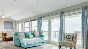 A living room with a teal sofa, patterned armchair, and a large window with curtains. - Beachfront home with beach access, deck, outdoor shower & washer/dryer (Galveston)