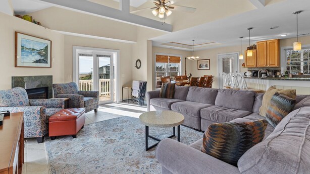 A spacious living room with a large gray sofa, a red ottoman, and patterned armchairs. A ceiling fan and pendant lights are visible. - Huge Ocean View Home w/ Elevator, Screened Porch, 2 Primary Suites & 3 Living Areas; Stroll to Beach (Oak Island)