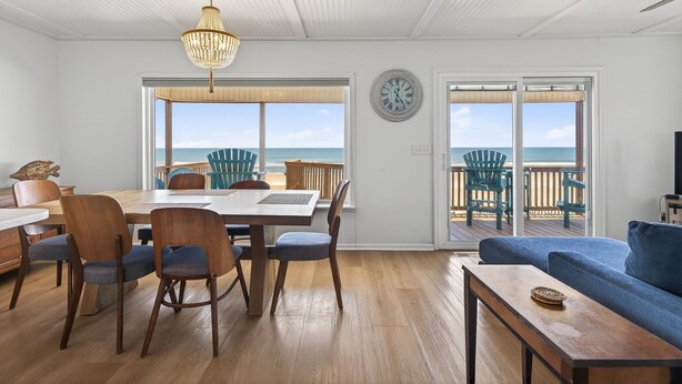 A dining area with a wooden table and chairs, a chandelier, and a view of the ocean through large windows.