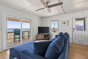 A living room with a large blue sofa, a ceiling fan, a television, and a view of the ocean through a sliding glass door.