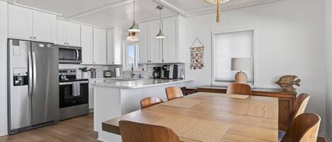 A modern kitchen with white cabinetry, stainless steel appliances, a wooden dining table, and a chandelier.