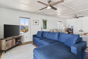 A living room with a blue sectional sofa, a wooden entertainment unit, a flat-screen TV, ceiling fans, and a view of the outdoors.