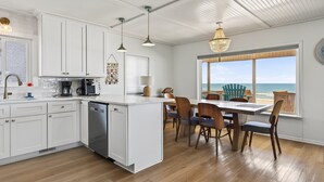 A modern kitchen with white cabinets, a central island, and a dining area with wooden chairs and a view of the ocean.