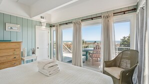 A bedroom with a bed, a wooden dresser, a wicker chair, and a view of the beach through large windows. - The Sanctuary -Premier Oceanfront Rental -Large Decks, Pet Friendly! (Folly Beach)