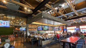 Amplia zona de bar con múltiples pantallas mostrando contenido deportivo, diversas opciones de asiento que incluyen taburetes y mesas, y un moderno diseño de techo industrial.