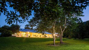 A well-lit house with a grassy lawn and trees in the foreground. - Luxury Home in Nature surrounded by Wallabies (Clifton Beach)